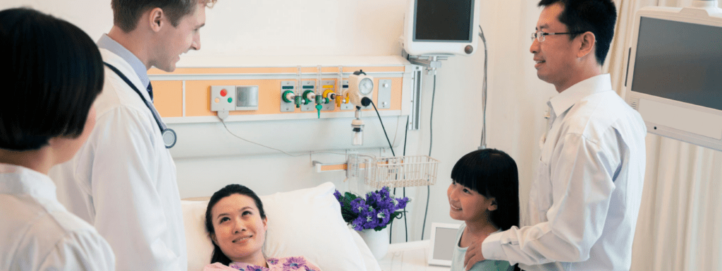 asian female patient in hospital bed surrounded by family and doctor 