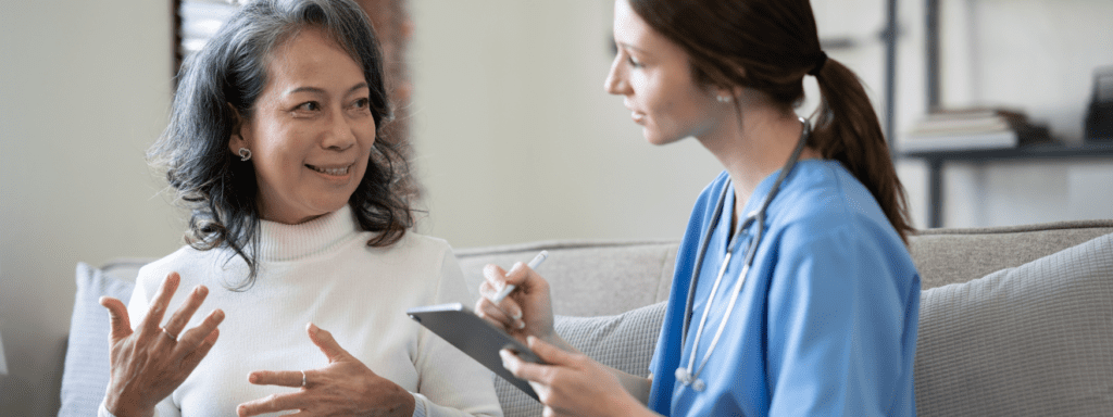 a woman with a stethoscope in a blue shirt taking notes talking to an older female patient in a white turtleneck shirt