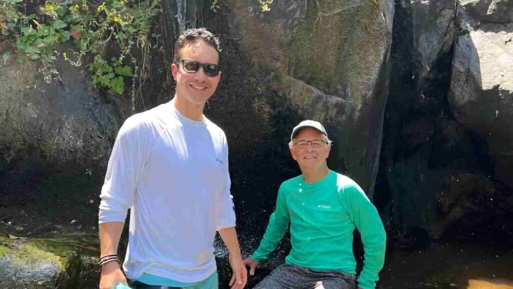 david brackett standing and his father sitting outside in front of large rocks wearing summer gear