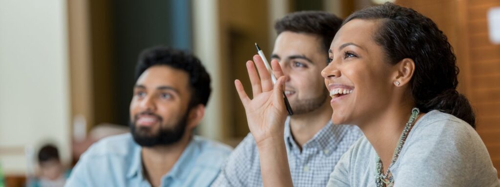 a closeup photo of a woman smiling and raising her hand in class with two men further behind her
