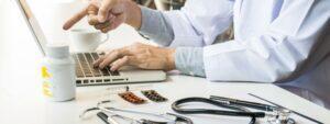 Doctor using a laptop at a desk with medical supplies and medication bottles nearby.
