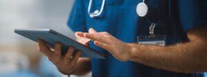 Healthcare worker in blue scrubs using a tablet, with one hand tapping the screen.