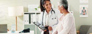 Doctor holding a tablet and smiling while speaking with an older patient in an exam room.