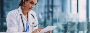 Clinician in a white coat reviewing information on a tablet beside a laptop in a medical setting.
